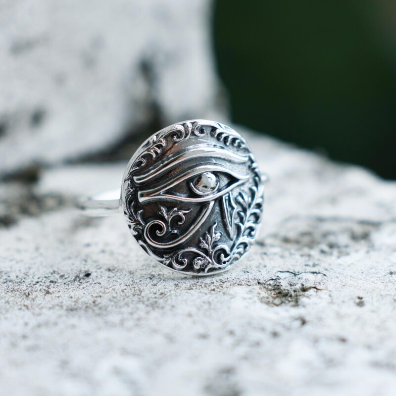 Close-up of a sterling silver Eye of Horus ring placed on a textured stone surface, showcasing the raised engraved symbol surrounded by ornamental floral detailing. The ancient Egyptian design is highlighted with soft natural shadows and polished oxidized finish.
