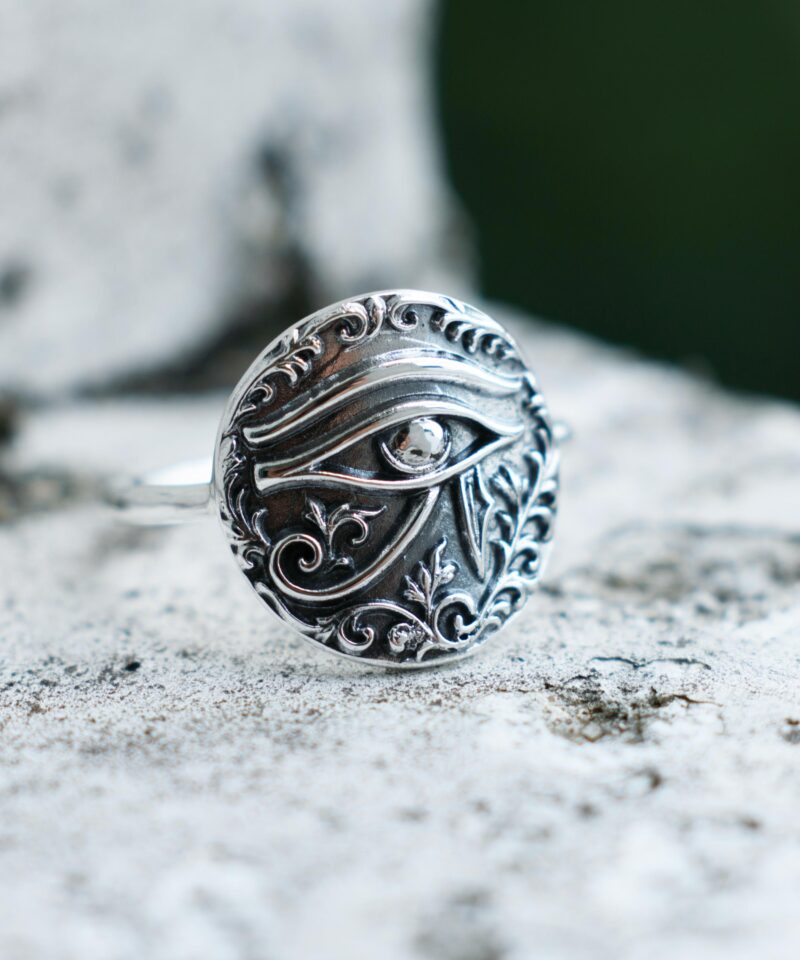 Close-up of a sterling silver Eye of Horus ring placed on a textured stone surface, showcasing the raised engraved symbol surrounded by ornamental floral detailing. The ancient Egyptian design is highlighted with soft natural shadows and polished oxidized finish.