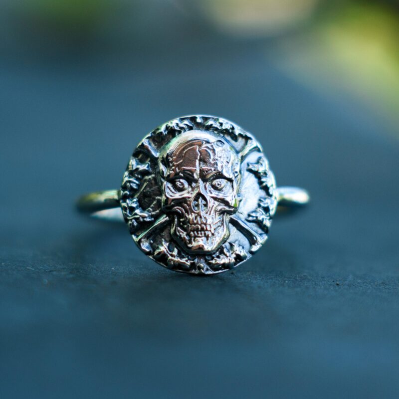 Front-facing image of a 925 sterling silver skull signet ring with high relief sculpting. The haunting skull design, set against a circular backdrop of bones, is perfect for alternative style lovers, rockers, or fans of occult and macabre symbolism. Its antique finish brings out every carved detail, turning this artisan ring into a wearable piece of art full of attitude and mystique.
