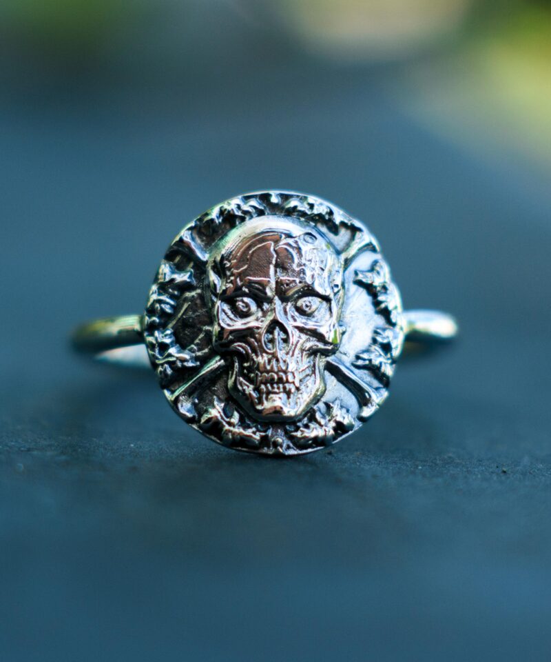 Front-facing image of a 925 sterling silver skull signet ring with high relief sculpting. The haunting skull design, set against a circular backdrop of bones, is perfect for alternative style lovers, rockers, or fans of occult and macabre symbolism. Its antique finish brings out every carved detail, turning this artisan ring into a wearable piece of art full of attitude and mystique.