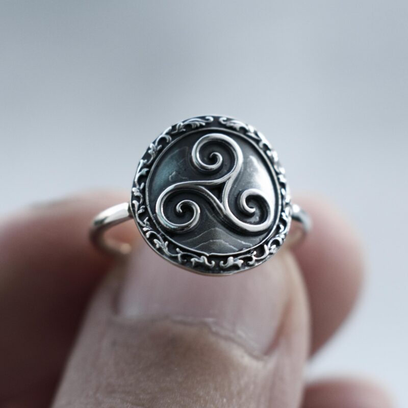 A close-up of the Triskelion ring worn on the finger, showing the detailed triple spiral Celtic symbol in the center. The oxidized floral border and high-polished sterling silver band contrast beautifully with natural daylight, emphasizing the symbolic craftsmanship.