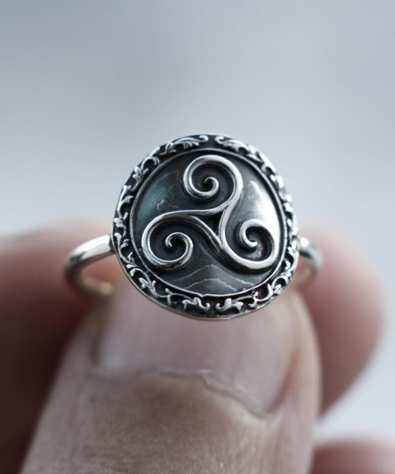 A close-up of the Triskelion ring worn on the finger, showing the detailed triple spiral Celtic symbol in the center. The oxidized floral border and high-polished sterling silver band contrast beautifully with natural daylight, emphasizing the symbolic craftsmanship.
