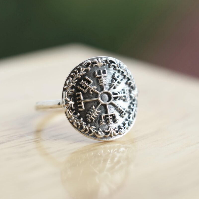 Sterling silver ring featuring a raised Vegvisir Viking compass symbol in the center, surrounded by an oxidized decorative border. Photographed on a warm wood surface with natural light, the image highlights the intricate Norse design and polished handmade craftsmanship.