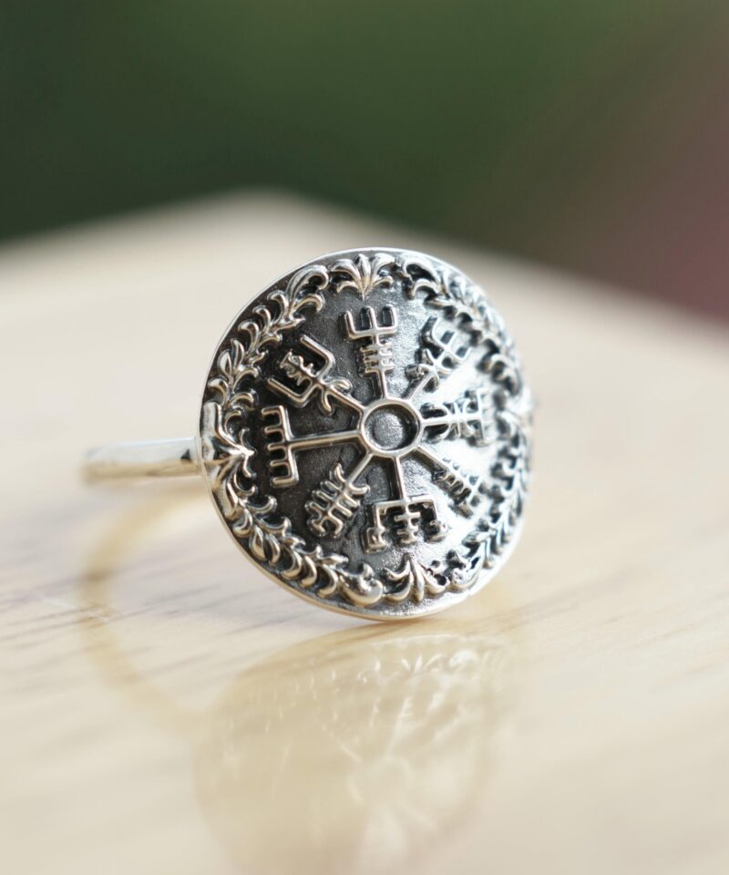 Sterling silver ring featuring a raised Vegvisir Viking compass symbol in the center, surrounded by an oxidized decorative border. Photographed on a warm wood surface with natural light, the image highlights the intricate Norse design and polished handmade craftsmanship.
