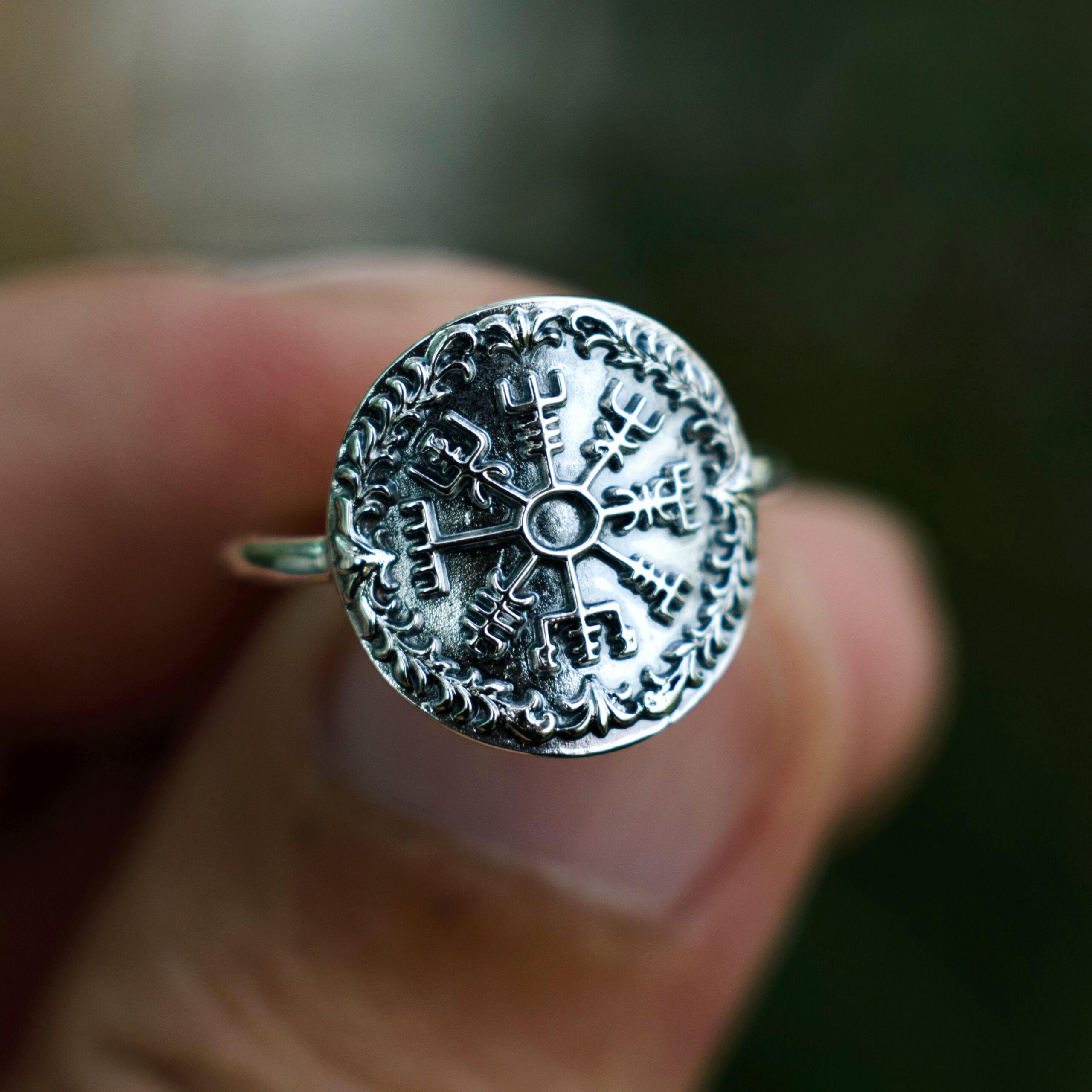 Close-up of a handmade silver Viking Vegvisir ring held between fingers, showing the intricate compass engraving surrounded by oxidized floral patterns. The photo uses natural outdoor lighting and a dark background to highlight the detail and spiritual depth of the symbol.